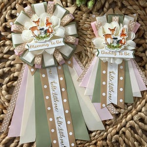 May include: Two baby shower ribbons with floral and woodland animal accents. One ribbon says "Mommy To Be" and the other says "Daddy To Be". Both have ribbons that say "hello little one". The ribbons are in shades of pink, green, and tan.
