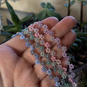 May include: Several beaded flower necklaces in pastel colors, including blue, pink, and green. The necklaces are made of small beads and are arranged in a floral pattern. The background is blurred, with green plants visible.