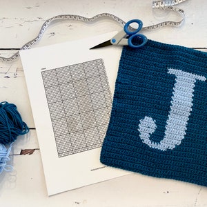 Peut inclure: Un carré crocheté bleu foncé avec un motif de lettre "J" bleu clair. Un graphique imprimé, des ciseaux, un mètre ruban et du fil sont également présents, suggérant un projet de création. L'image est sur une surface en bois blanc.