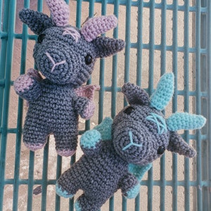 K&ouml;nnte beinhalten: Zwei graue geh&auml;kelte Drachen-Pl&uuml;schtiere mit blauen und rosa Akzenten. Die Drachen sitzen auf einer blauen und wei&szlig;en Gitteroberfl&auml;che.
