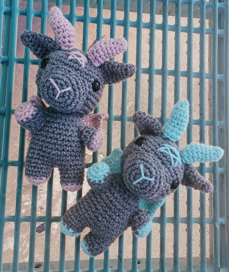 K&ouml;nnte beinhalten: Zwei graue geh&auml;kelte Drachen-Pl&uuml;schtiere mit blauen und rosa Akzenten. Die Drachen sitzen auf einer blauen und wei&szlig;en Gitteroberfl&auml;che.