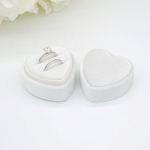 Op de afbeelding: Twee zilveren ringen met heldere edelstenen in een open, witte, hartvormige fluwelen doos. Een bijpassende gesloten doos staat ernaast. Beide liggen op een witte ondergrond. Voor een speciale gelegenheid.
