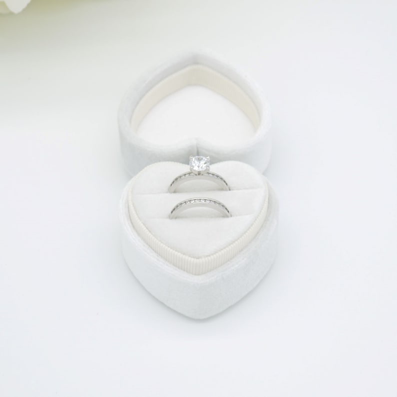 Op de afbeelding: Een witte, hartvormige ringendoos met twee zilveren ringen. De bovenste ring heeft een grote, heldere edelsteen. De onderste ring is een band met kleine, heldere edelstenen. De doos is open en toont de ringen.