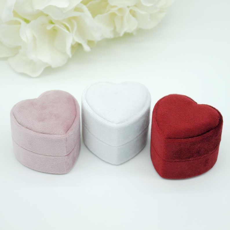 Op de afbeelding: Drie hartvormige ringdozen in roze, witte en rode fluweel. De dozen zijn gesloten, met een zachte, pluche textuur. De achtergrond is wit, met een vage bloemstuk bovenaan.