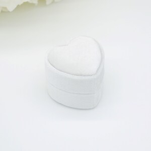 Op de afbeelding: Een witte, hartvormige ringdoos. De doos is gemaakt van een zacht, getextureerd materiaal en heeft een afgeronde vorm. De bovenkant van de doos is ook hartvormig, waardoor een samenhangend ontwerp ontstaat. De doos staat tegen een witte achtergrond.