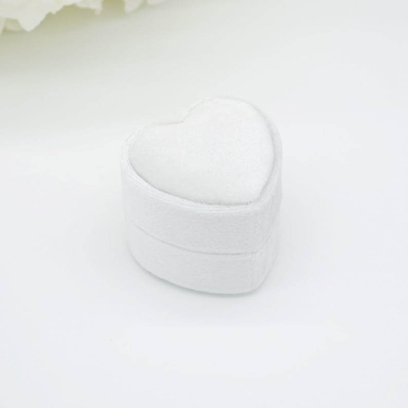 Op de afbeelding: Een witte, hartvormige ringdoos. De doos is gemaakt van een zacht, getextureerd materiaal en heeft een afgeronde vorm. De bovenkant van de doos is ook hartvormig, waardoor een samenhangend ontwerp ontstaat. De doos staat tegen een witte achtergrond.