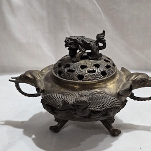 Puede incluir: Antiguo quemador de incienso de bronce con detalles intrincados. El quemador presenta una tapa decorativa con una pequeña figura de león en la parte superior. El cuerpo está adornado con motivos de aves y dragones, y tiene asas a cada lado. El quemador se apoya en tres patas.