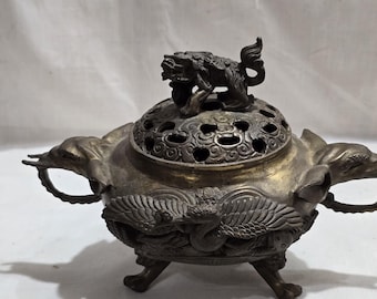 Puede incluir: Antiguo quemador de incienso de bronce con detalles intrincados. El quemador presenta una tapa decorativa con una pequeña figura de león en la parte superior. El cuerpo está adornado con motivos de aves y dragones, y tiene asas a cada lado. El quemador se apoya en tres patas.
