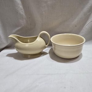 May include: A cream-colored ceramic gravy boat with a handle and a matching bowl. The gravy boat has a curved spout and sits on a small base. The bowl is round with a slightly flared rim and horizontal ridges.