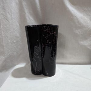 Puede incluir: Un jarrón de cerámica negro con una forma curva única. El jarrón presenta un acabado brillante y está adornado con delicados diseños de líneas abstractas rosas. El jarrón es un objeto decorativo.