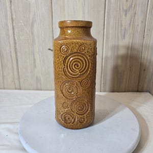 Puede incluir: Un jarrón de cerámica marrón con forma rectangular y superficie texturizada. El jarrón presenta una serie de diseños en espiral. El jarrón es un elemento decorativo, adecuado para contener flores o como pieza independiente.