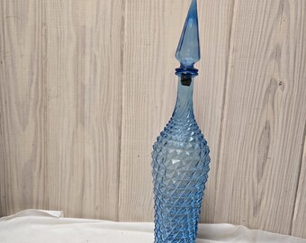 Puede incluir: Un decantador de vidrio azul claro alto con un patrón de diamantes texturizado en el cuerpo. El recipiente tiene un cuello esbelto y espiralado, rematado con un tapón de vidrio a juego, cónico y puntiagudo, que encaja en la abertura de la botella.