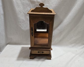 Puede incluir: Un pequeño joyero de madera con puerta de espejo y un pequeño cajón. El armario es de color marrón cálido y presenta herrajes decorativos. La parte superior del armario tiene un diseño curvo. El armario está diseñado para colocarse sobre una superficie plana.