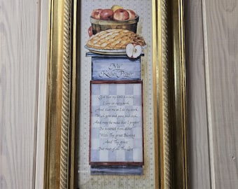 Puede incluir: Un cuadro rectangular con marco dorado que muestra una cesta de manzanas y un pastel sobre el texto My Kitchen Prayer. La impresión incluye un poema sobre bendiciones en la cocina sobre un fondo de cuadros en azul claro y blanco.