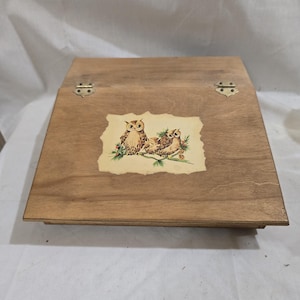May include: A wooden box with a hinged lid and two brass hinges. The box has a decorative design of three owls on a branch with pine needles and berries. The box is a light brown color.