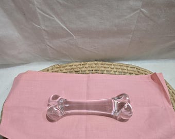 Puede incluir: Un objeto transparente en forma de hueso de cristal, probablemente decorativo, descansa sobre una tela rosa. El hueso tiene extremos redondeados y una superficie lisa. La tela está colocada sobre una estera redonda tejida.