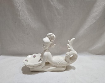 Puede incluir: Figura de sirena de cerámica blanca, reclinada con un plato en forma de concha. La sirena tiene escamas detalladas y una cola fluida. El plato tiene pequeños acentos florales morados. La figura mide unos 20 cm de largo.