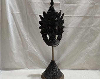 Puede incluir: Una escultura negra de la cabeza de una deidad con un tocado adornado, montada sobre un soporte de color latón. El soporte tiene una base cónica con patrones detallados y una base circular. La escultura mide aproximadamente 30 cm de altura.