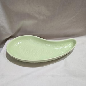 Puede incluir: Un plato de cerámica verde pálido en forma de lágrima. El plato tiene un borde ligeramente elevado y un delicado estampado floral en relieve en su superficie. El diseño es simple y elegante, adecuado para servir aperitivos o porciones pequeñas.