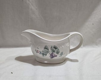 Puede incluir: Salsera de cerámica blanca con asa y boquilla. El recipiente está decorado con un motivo floral con hojas verdes y racimos de uvas moradas. La salsera se asienta sobre una pequeña base redonda.