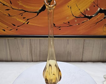 Puede incluir: Un florero de vidrio color ámbar, alto y esbelto, con cuello estrecho y base bulbosa con nervaduras verticales. El florero presenta un borde acampanado de forma orgánica y se apoya sobre una base circular con un patrón de pétalos.