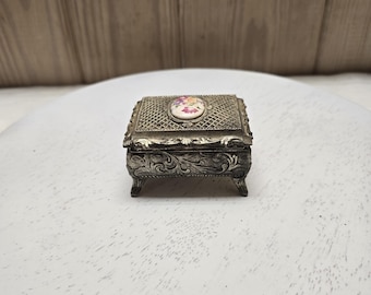 Puede incluir: Un pequeño joyero rectangular de metal plateado con patrones de volutas en relieve en los laterales y cuatro patas decorativas cortas. La tapa presenta un diseño de rejilla texturizada con una incrustación floral ovalada central.
