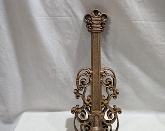 Puede incluir: Un candelabro de pared de color bronce en forma de violín. El candelabro tiene adornos de volutas y un diseño detallado. La copa de la vela está en la parte inferior y el diseño general es simétrico.