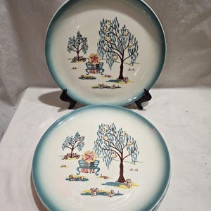May include: Two vintage ceramic plates with a teal rim and a scenic illustration. The plates feature a park scene with trees, a bench, and floral accents in shades of blue, brown, pink, and yellow. The plates are displayed on a black stand.