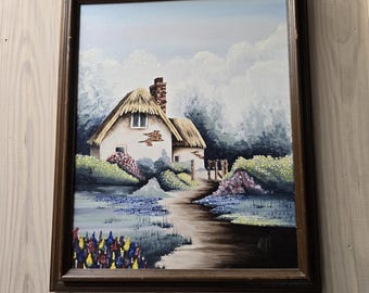 Puede incluir: Una pintura al óleo enmarcada que representa una casa de campo blanca con techo de paja, una chimenea de ladrillo y un camino de tierra que conduce a la entrada. La escena incluye flores coloridas en primer plano, arbustos y cielo azul.