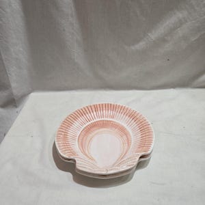 Puede incluir: Un plato de cerámica con forma de concha, en tonos rosa claro y blanco. El plato tiene un borde festoneado y una superficie texturizada, ideal para servir pequeñas porciones o como elemento decorativo.
