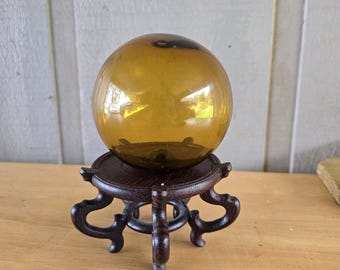 Puede incluir: Una esfera de cristal de color ámbar, lisa y translúcida, que descansa sobre un soporte de madera de color marrón oscuro. El soporte cuenta con una plataforma superior circular sostenida por cuatro patas curvas y ornamentadas.