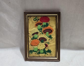 Puede incluir: Obra de arte bordada con un grupo de setas coloridas en tonos rojos, naranjas, amarillos y marrones. La obra está enmarcada en un marco de madera oscura y sobre un fondo de tela amarilla. Se ve una pequeña mariposa verde.