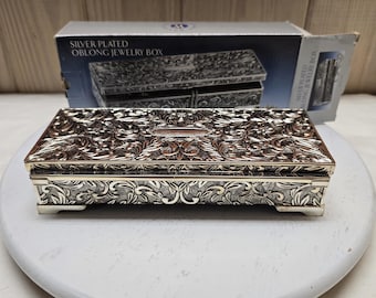 Puede incluir: Un joyero oblongo chapado en plata con un intrincado patrón floral y de hojas en relieve en la tapa y los laterales. La caja incluye una placa de grabado rectangular central en la tapa y se apoya sobre cuatro patas pequeñas. Caja original incluida.