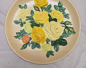 Puede incluir: Un plato decorativo redondo de color crema con un diseño floral en relieve tridimensional. El centro muestra un grupo de rosas amarillas y amarillo pálido con hojas verdes y un pequeño capullo de rosa naranja sobre el plato liso.