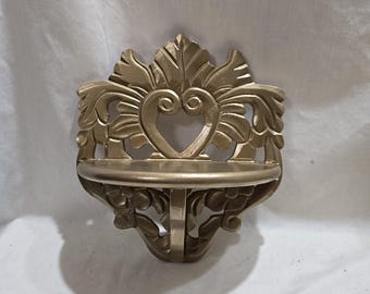 Puede incluir: Estante de pared dorado con detalles ornamentales en forma de hojas. El estante presenta un recorte en forma de corazón en el centro y un pequeño estante redondo. El diseño es simétrico y decorativo.
