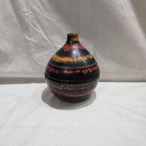 May include: A decorative ceramic jar with a lid. The jar is dark with horizontal bands of orange, red, and brown. The jar has a rounded body and a narrow neck. The lid fits snugly on top of the jar.