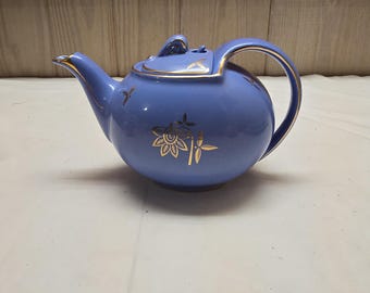 Puede incluir: Una tetera de cerámica azul bígaro con un motivo floral y de bambú dorado en el cuerpo y la tapa. La tetera tiene un asa curva, un pico puntiagudo y un borde dorado a lo largo del pico, el asa y la tapa, con un pequeño remate en la parte superior.