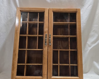 Puede incluir: Un mueble de madera con dos puertas de cristal. El mueble tiene un acabado marrón claro y presenta múltiples compartimentos pequeños en el interior. Se ven tiradores decorativos de metal en las puertas. El mueble está diseñado para exhibición o almacenamiento.