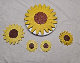 Puede incluir: Un conjunto de cinco placas decorativas de pared con forma de girasol en varios tamaños. Cada pieza presenta pétalos de color amarillo brillante que irradian desde un centro marrón oscuro texturizado. El girasol más grande está montado sobre una base circular blanca, mientras que los cuatro más pequeños están dispuestos alrededor, mostrando un diseño floral consistente con crestas detalladas en los pétalos.