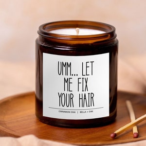 May include: A brown glass candle jar with a white label that reads "Umm... Let Me Fix Your Hair". The candle is on a wooden tray and has a white wick.