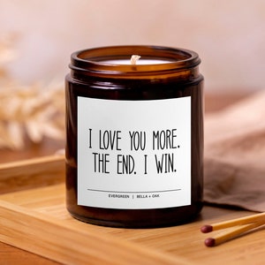 May include: A brown glass candle jar with a white label that reads "I LOVE YOU MORE. THE END. I WIN." The candle is on a wooden tray with two matchsticks.