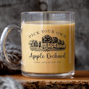 May include: A clear glass mug with a black and white graphic of an apple orchard with the text "Pick Your Own Apple Orchard - Cider - Apple Butter - Pies". The mug is filled with a light brown liquid.