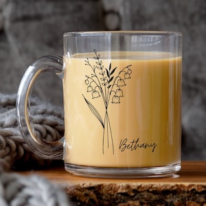 May include: A clear glass mug with a black line drawing of lily of the valley flowers and the name "Bethany" printed on the side. The mug is filled with a light brown beverage.