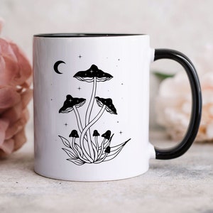 May include: White ceramic mug with a black rim and handle. The mug features a black and white illustration of mushrooms with a crescent moon and stars in the background.