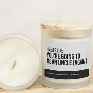May include: A frosted glass candle jar with a wooden lid. The candle is white and has a label that reads "Smells Like You're Going To Be An Uncle (Again)". The label also includes the text "Hand Poured | Cashmere Vanilla | Bella & Oak".