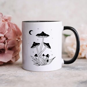 May include: A white ceramic mug with a black handle and interior, featuring a black line art illustration of a cluster of mushrooms, a crescent moon, and small stars on the front surface. The design is centered on the exterior of the mug.
