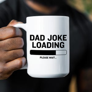 May include: A white ceramic mug featuring the black text DAD JOKE LOADING, a black progress bar graphic, and the text PLEASE WAIT below it. The mug has a smooth finish, a curved handle on the left side, and a cylindrical shape for holding beverages.
