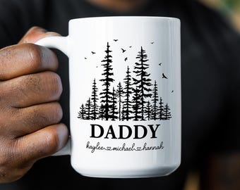 May include: A white ceramic mug featuring a black graphic of a pine forest with birds flying above. The word DADDY is printed in a bold black font below the trees, followed by the names kaylee, michael, and hannah separated by heart icons.