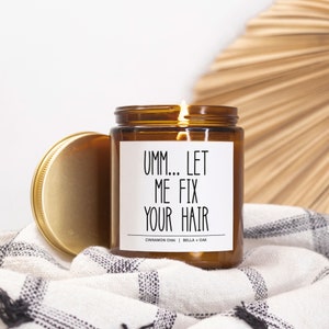 May include: A brown glass candle with a gold lid. The candle has a white label with the text "Umm... Let Me Fix Your Hair" and the scent "Cinnamon Chai | Bella + Oak".