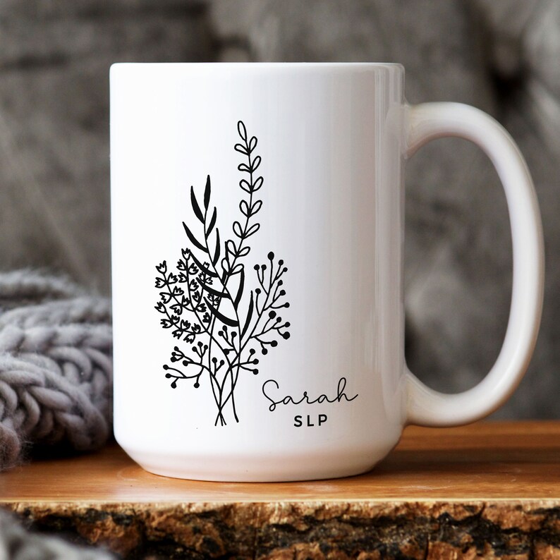 May include: White ceramic mug featuring a black floral design with the name "Sarah" and the initials "SLP" in a delicate script. The mug has a large handle and is set on a wooden surface.